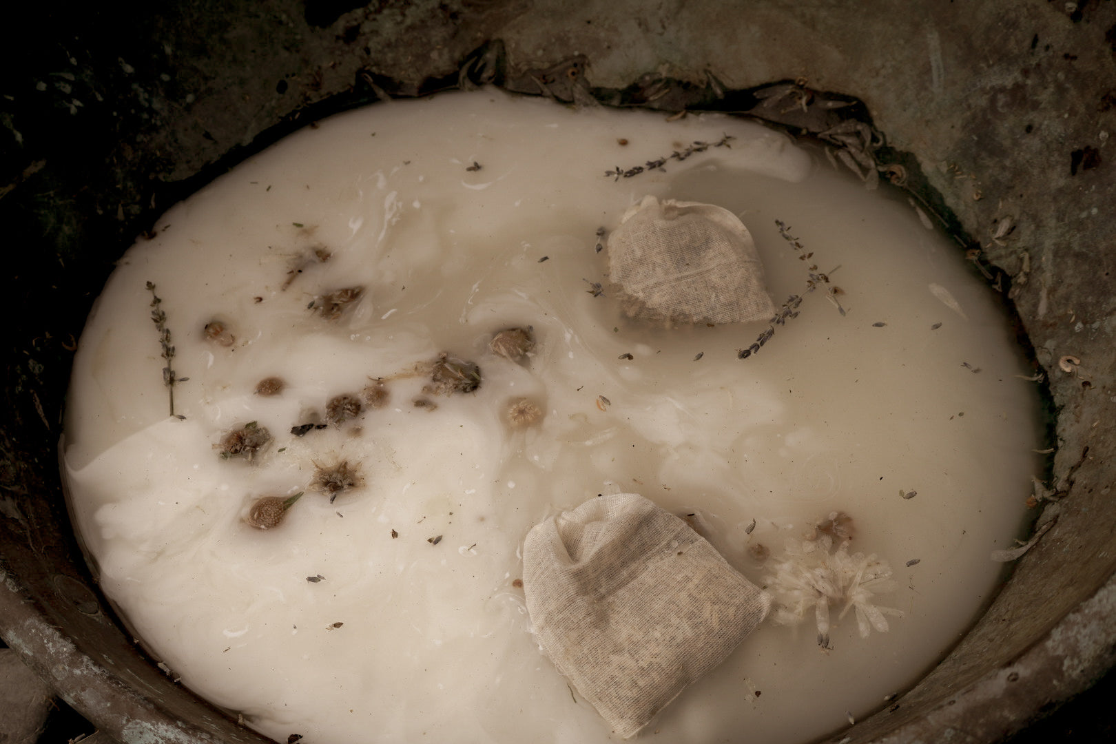 Rosemary and mint herbal bath bags floating in a pale milky bath with botanicals